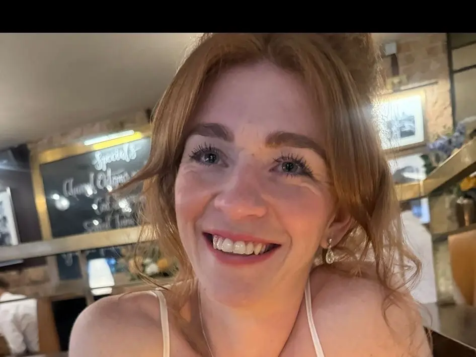 Housewife_kirst smiles while holding her phone up, showing her face, neck, and shoulders with her light brown hair down.