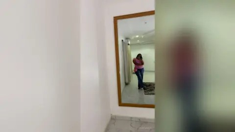 Woman in pink top and blue jeans standing in a bright modern room reflecting in a large wall mirror.