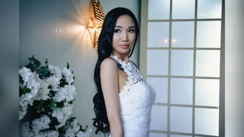 Elegant woman in a white lace wedding dress posing in a bright, minimalist room with floral decor.