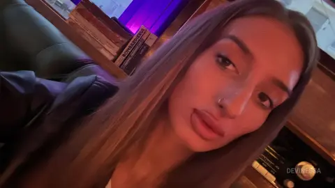 DeviNessa looks into the camera with long brown hair and a nose ring while sitting in a room with purple lighting.