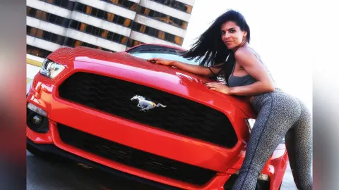 DrellaWolf — Smiling woman in grey athletic wear leaning on a red Ford Mustang in a bright urban setting.