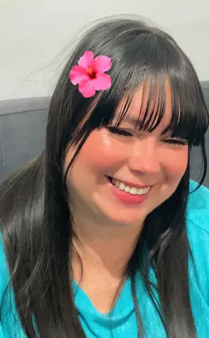 Smiling woman with dark hair and a pink hibiscus flower in a bright teal shirt in a cozy indoor setting.