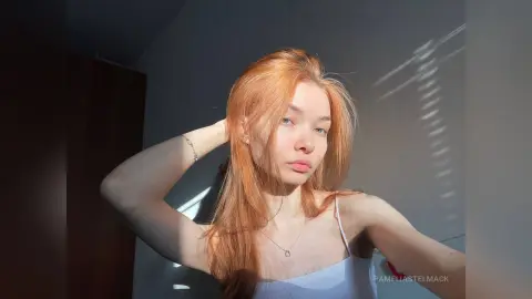 Redheaded woman in a white tank top posing in soft sunlight against a shadowed wall.
