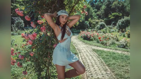 AlessiaCrush poses in a white lace romper and headscarf while standing next to pink rose bushes in a sunny garden.