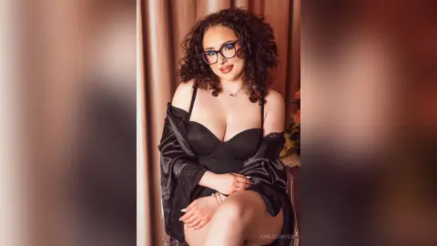 Curvy woman with curly hair in a black bodysuit and glasses posing glamorously in a warm indoor setting.