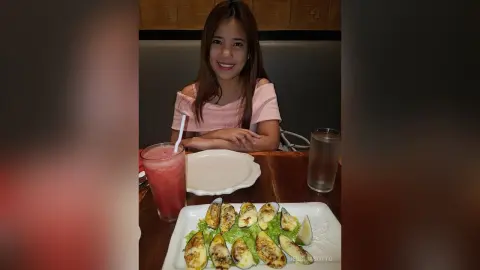 Smiling woman in a pink off-the-shoulder top enjoying a seafood meal and refreshing drink at a restaurant.
