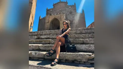 AnaMathers sits on stone steps wearing a black dress and sunglasses against a large stone building.