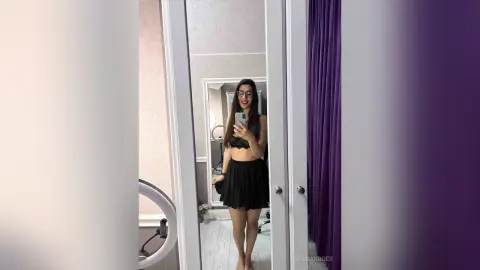 Smiling young woman in a black crop top and skirt taking a mirror selfie in a bright modern room.