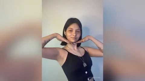 JacquiKozma poses with her hands under her chin, wearing a black tank top against a plain light wall.