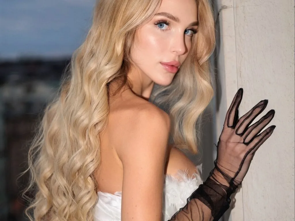 LuluBianco poses in a white feathered outfit and sheer black gloves with her long blonde hair flowing over her shoulder.
