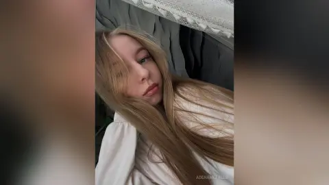 AdenaMcfalls poses with her long blonde hair draped over her shoulder, wearing a white top against a grey background.