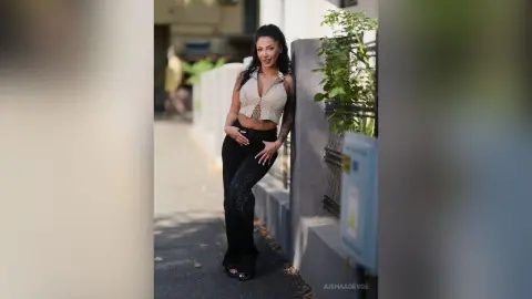 Stylish woman in a beige crop top and black trousers leaning against a wall on a sunny urban street.