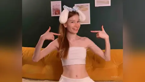 Smiling young woman in a white crop top and bunny ears sitting on a yellow sofa in a cozy room.