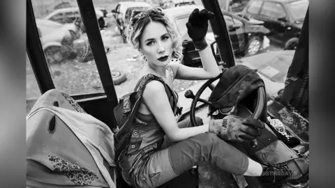 ChristineDavis poses in a car wearing overalls and black gloves, looking back over her shoulder with blonde hair and dark lipstick.