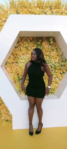 Stylish woman in a black cocktail dress standing before a decorative yellow floral hexagon wall.
