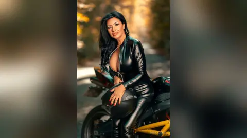 MissyJolie poses in a black leather bodysuit while sitting on a motorcycle against a soft, golden outdoor background.