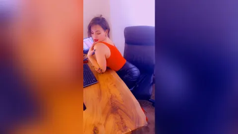 Selena — Selena sits at a wooden desk in a red tank top and black leather pants, leaning forward with her chin on her hand.