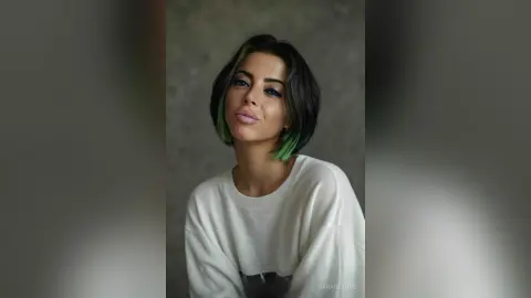 Young woman with short dark hair and green highlights wearing a white sweatshirt in a soft, moody portrait.