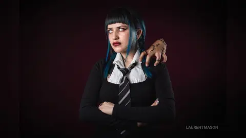 Goth woman with blue hair and black outfit posing with a moody expression against a dark background.