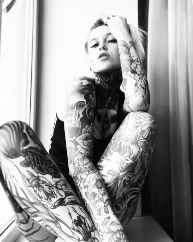 Black and white portrait of a tattooed woman in a black bodysuit sitting by a window in a moody setting.
