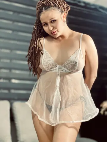 Woman with long dreadlocks wearing a white lace sheer babydoll nightgown in a moody indoor setting.