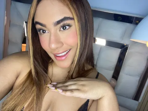 Smiling woman with long brown hair and glamorous makeup wearing black lace lingerie in a bright bedroom.