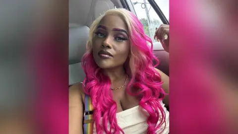 BossyBella — BossyBella poses in a car with long, wavy pink and blonde hair, wearing a white top and a gold chain necklace.