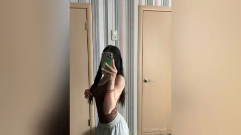 Young woman in a brown tank top taking a mirror selfie in a minimalist room with neutral tones.