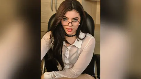 Young woman with long dark hair and glasses wearing a white button-down shirt in a cozy indoor setting.