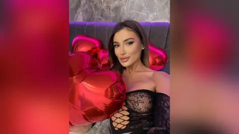 Glamorous woman in a black lace outfit holding red heart balloons in a moody, romantic setting.