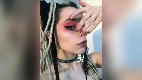 Woman with long blonde dreadlocks and pink eye makeup wearing a black studded choker in a soft-lit setting.