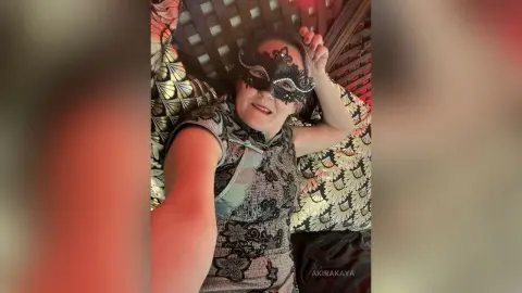 Smiling woman in a patterned qipao dress and black lace masquerade mask relaxing in a cozy setting.