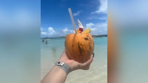Hand holding a fresh coconut cocktail with a straw on a sunny tropical beach with turquoise ocean water.