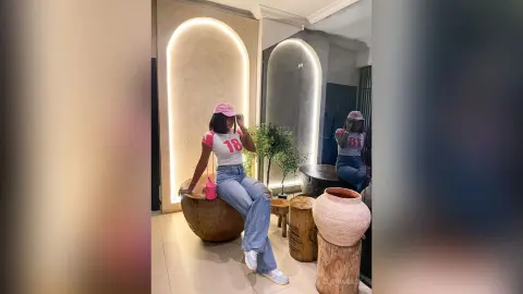 Young woman in a white graphic t-shirt and blue jeans posing in a modern, minimalist interior with warm lighting.