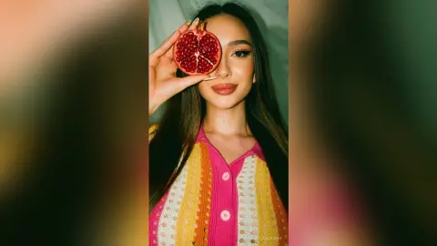 Young woman in a colorful knit cardigan holding a sliced pomegranate over her eye in a bright, playful mood.