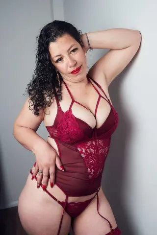 Curvy woman in red lace bodysuit and lingerie posing against a grey wall with a confident mood.