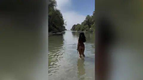A woman in a bikini wading through a tranquil tropical river surrounded by lush green palm trees.