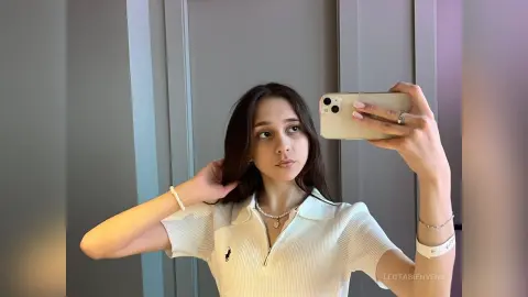 Young woman in a white ribbed polo shirt taking a mirror selfie in a modern minimalist room.