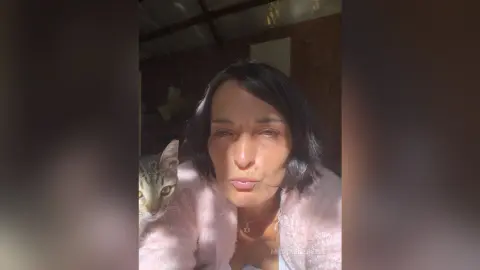 MichaelaBelle pouts her lips while wearing a soft pink fuzzy sweater next to a tabby cat.