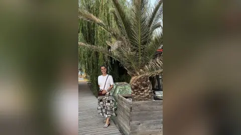 Smiling woman in a white top and black patterned skirt standing by palm trees in a sunny outdoor setting.