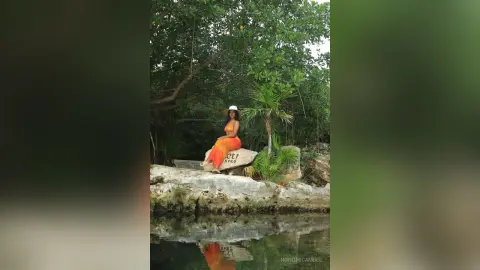 NohomiCambel sits on a rock near a pool of water wearing an orange sundress and a white hat against a backdrop of green trees.