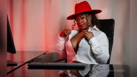 Confident woman in a white shirt and red hat sitting at a desk in a moody, red-lit office setting.