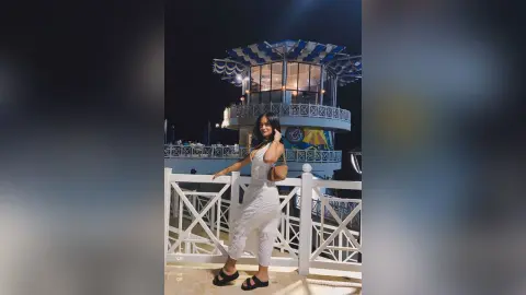 Young woman in a white sleeveless outfit posing on a balcony at a lit rooftop lounge at night.