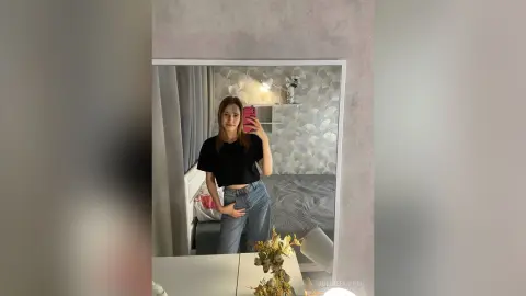 Young woman in a black t-shirt and blue jeans taking a mirror selfie in a cozy, modern bedroom.