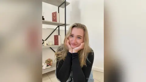 Smiling blonde woman in a black long-sleeve shirt relaxing in a bright, minimalist room with bookshelves.