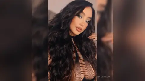 Glamorous woman with long wavy dark hair wearing a black sheer mesh cutout top in a soft indoor setting.