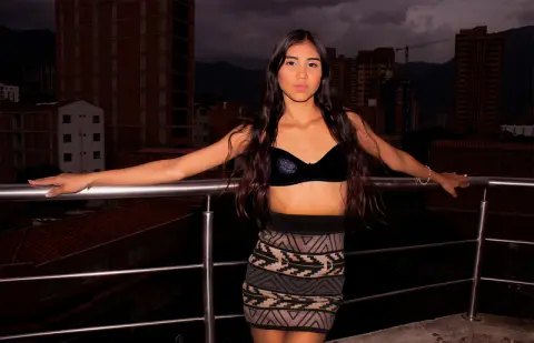 Young woman in a black crop top and patterned mini skirt posing on a city rooftop at night.