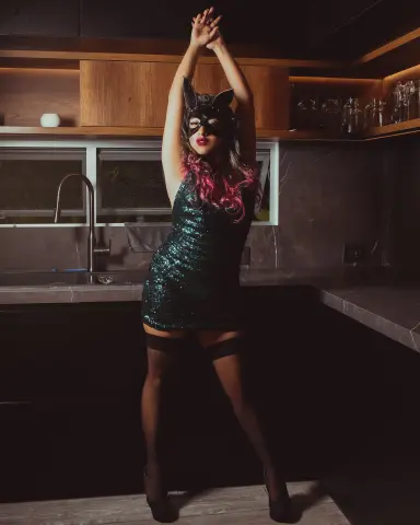 Glamorous woman in a sparkly green sequin dress and cat mask posing in a moody, modern kitchen setting.