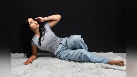 KaiaSinclair lies on a white shag rug wearing a grey t-shirt and light blue jeans, looking up with her hand in her long dark hair.