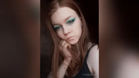 Young woman with teal eyeshadow and long red hair posing in a dark setting with a moody aesthetic.